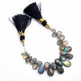 Labradorite faceted pear gemstone bead strand with silk tassels, dark grey with spectacular blue-green flash color, arranged on white background, jewelry making supply bead strand